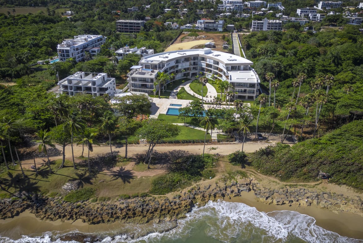 Aerial view of a modern curved resort complex with pools and palm trees on a tropical coastline.