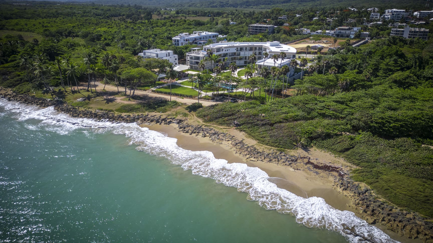 Aerial view of a modern resort complex with white buildings, pools, and palm trees on a tropical coastline.
