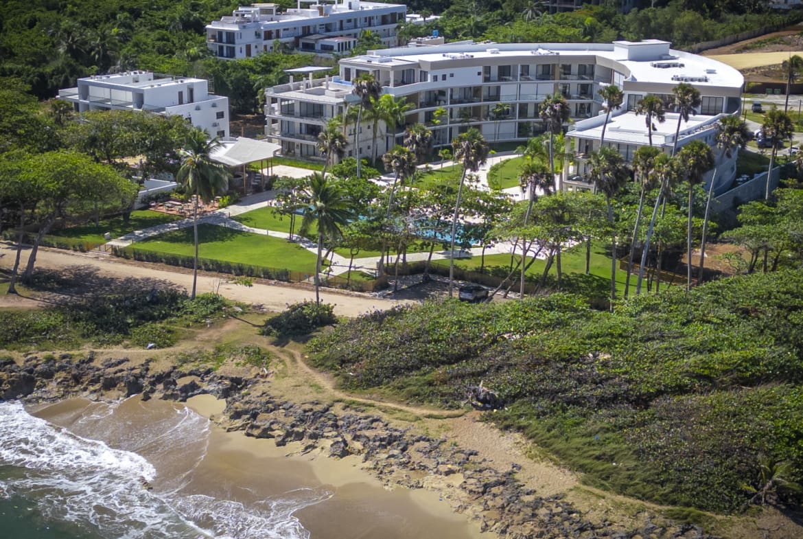 Aerial view of a modern resort hotel with a pool and lush green landscaping by the ocean.
