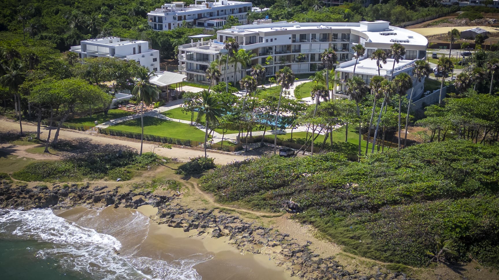 Aerial view of a modern resort hotel with a pool and lush green landscaping by the ocean.