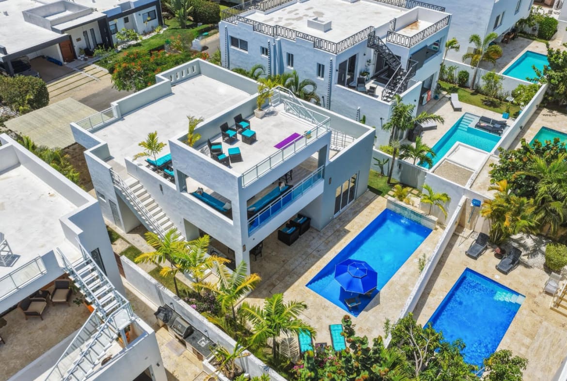 Modern white villas with blue swimming pools and palm trees in a tropical setting.