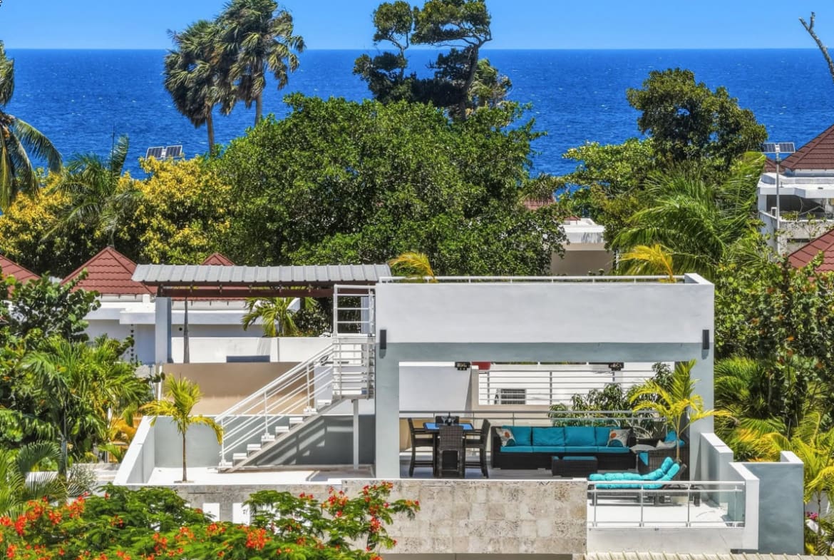 Modern rooftop terrace with ocean view, turquoise seating, and lush tropical foliage.