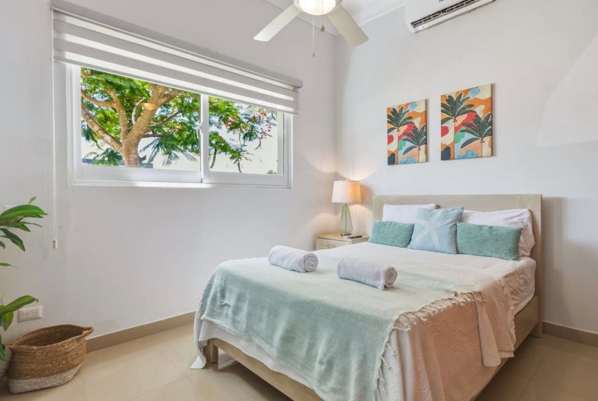 Bright bedroom with a queen bed, tropical artwork, and a window view of palm trees.
