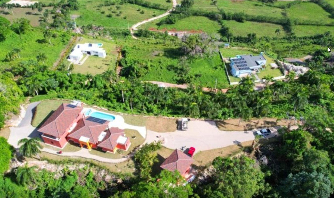 Aerial view of a sprawling red villa with a pool, surrounded by lush greenery and modern homes.
