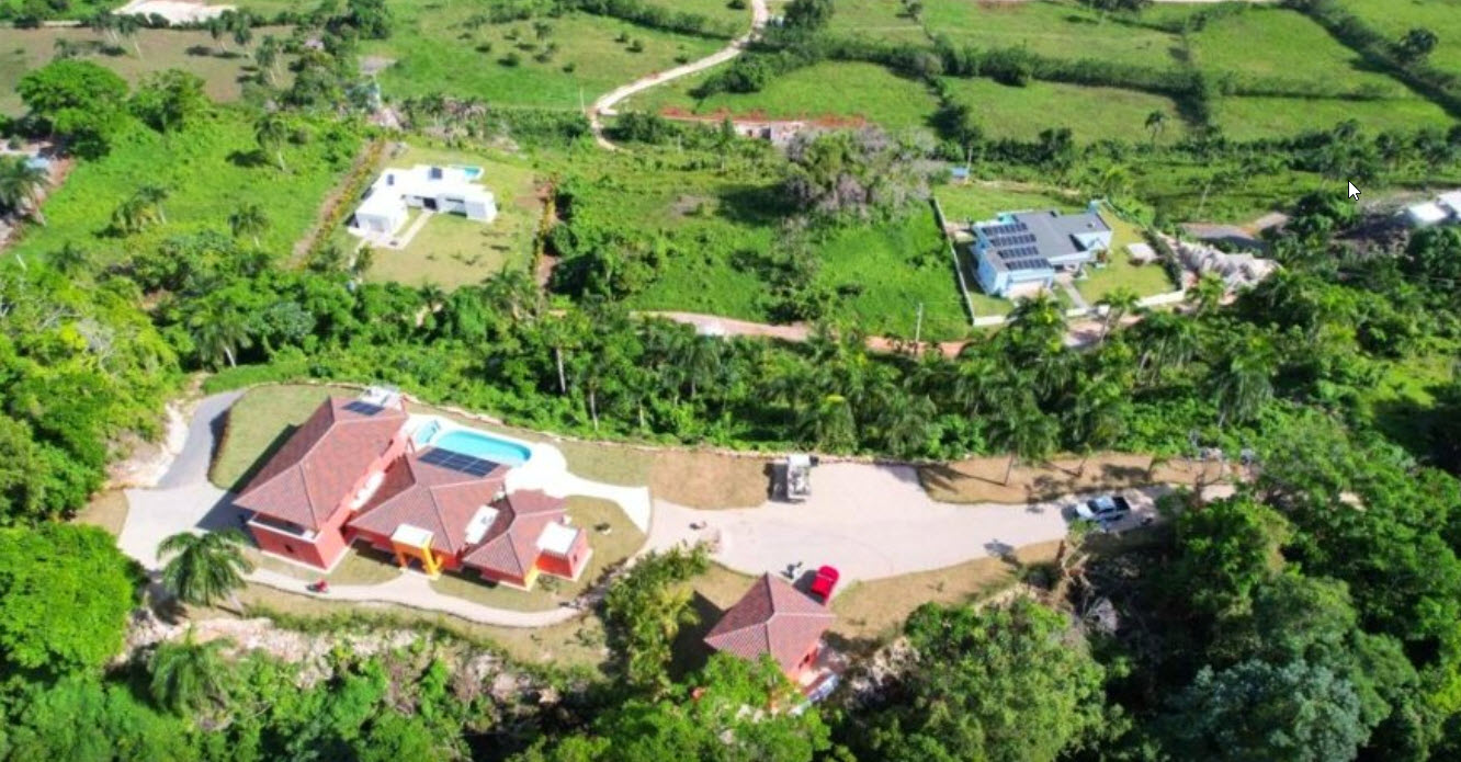 Aerial view of a sprawling red villa with a pool, surrounded by lush greenery and modern homes.