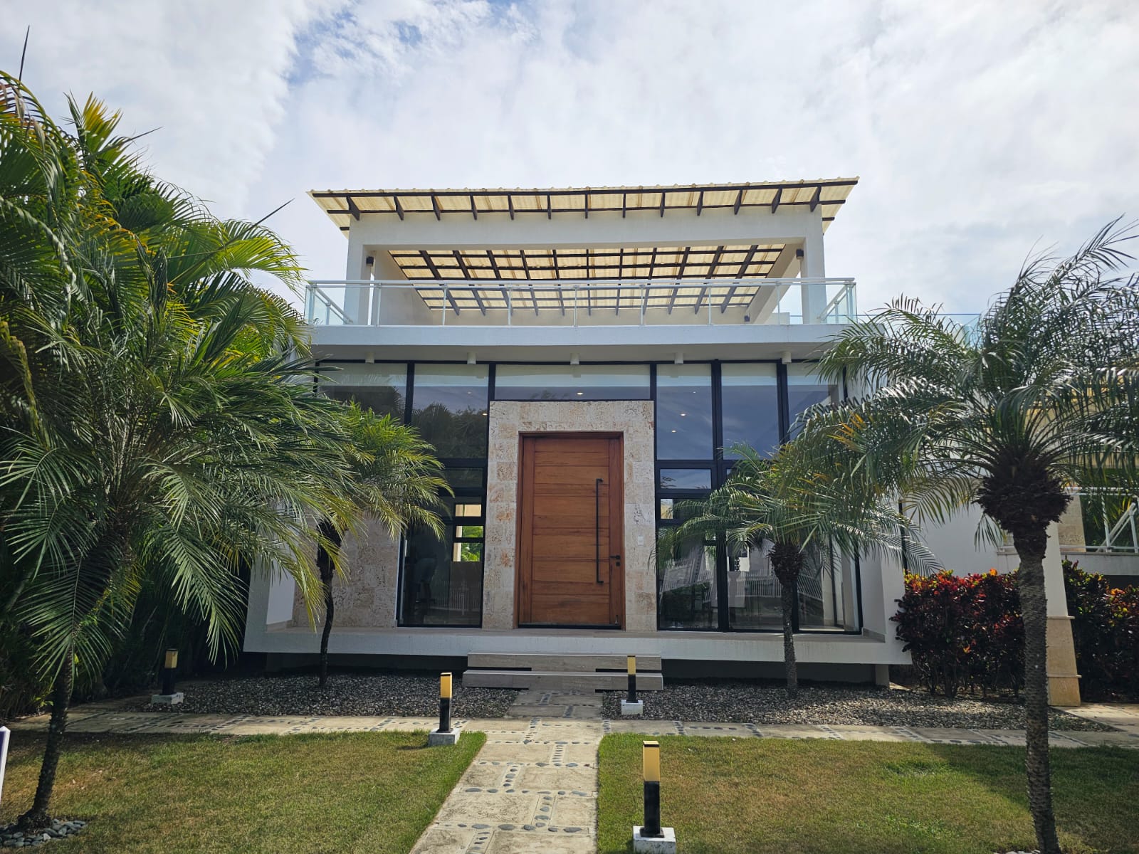 Modern white house with large glass windows and a wooden door, surrounded by palm trees.