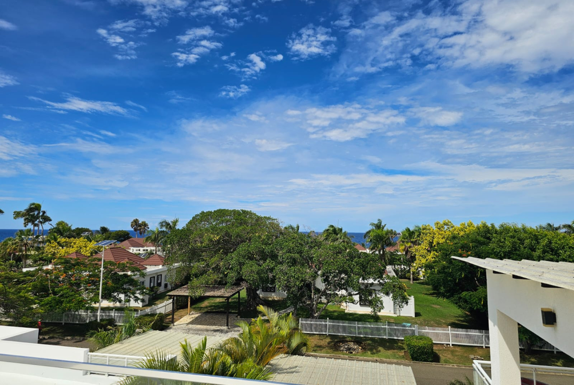 Tropical resort with white buildings, lush trees, and blue ocean under a cloudy sky.