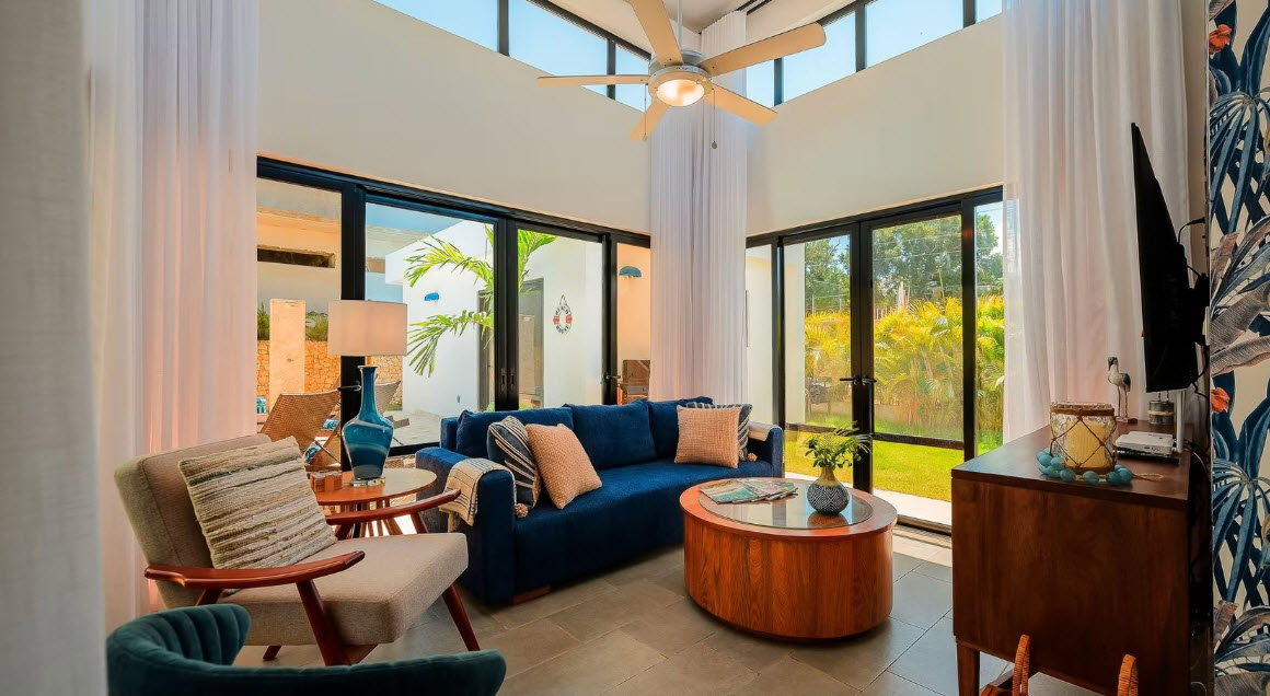 Modern living room with blue sofa, large windows, and ceiling fan overlooking a garden.