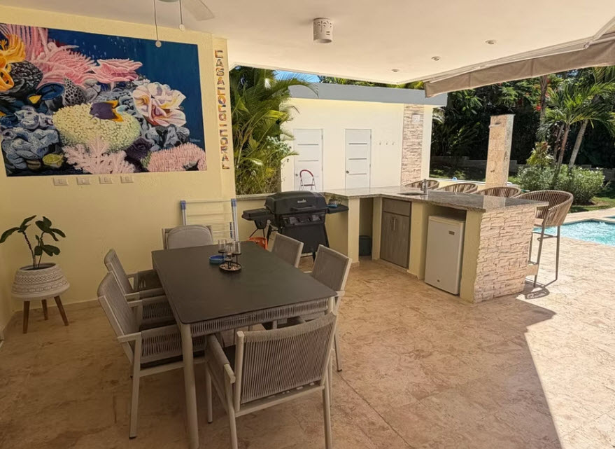 Outdoor kitchen with dining table, grill, and bar seating by a pool.