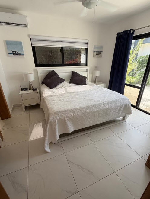Modern white bedroom with king-size bed, nightstands, and large window overlooking lush greenery.
