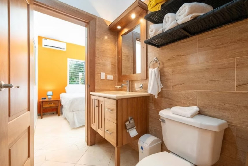 Modern bathroom with wooden vanity, tiled walls, and view of bedroom with orange wall.
