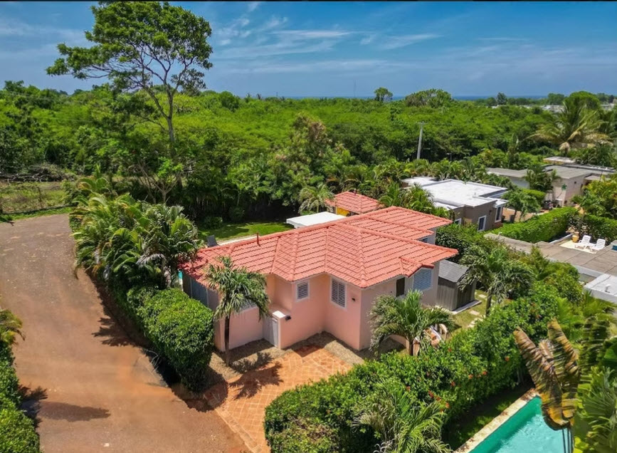 Aerial view of a pink villa with a red tile roof, lush greenery, and a swimming pool.