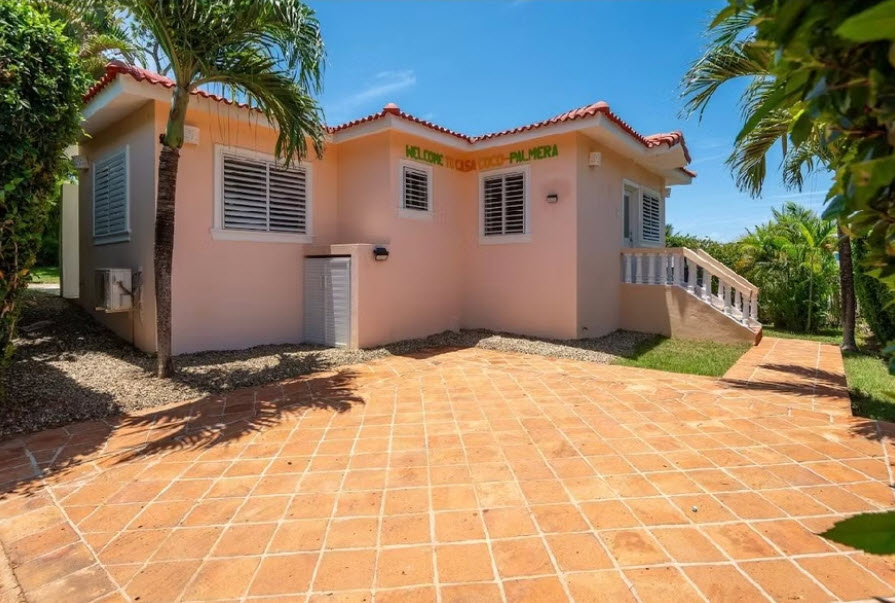 Pink villa with "Welcome to Coco Palmira" sign, palm trees, and tiled driveway.