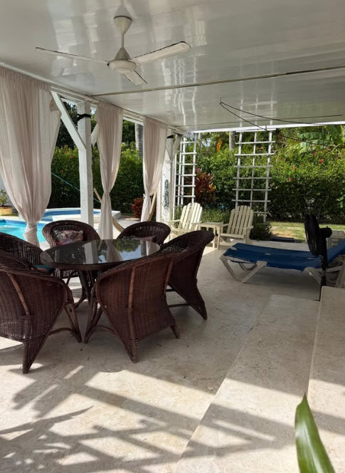 Covered patio with wicker furniture, lounge chairs, and a pool in a tropical garden.