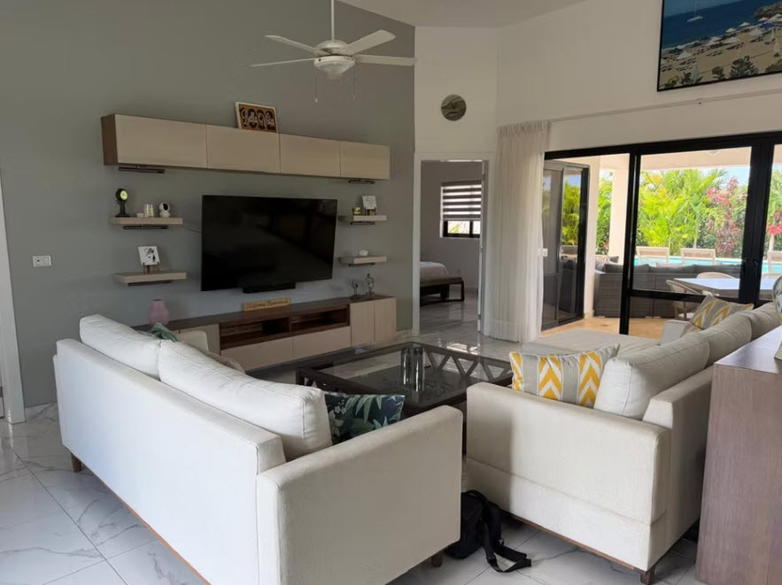 Modern living room with white sofas, large TV, and view of tropical pool area