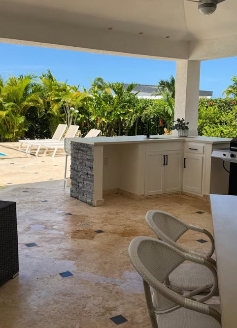 Outdoor kitchen with grill, sink, and seating area surrounded by tropical plants.