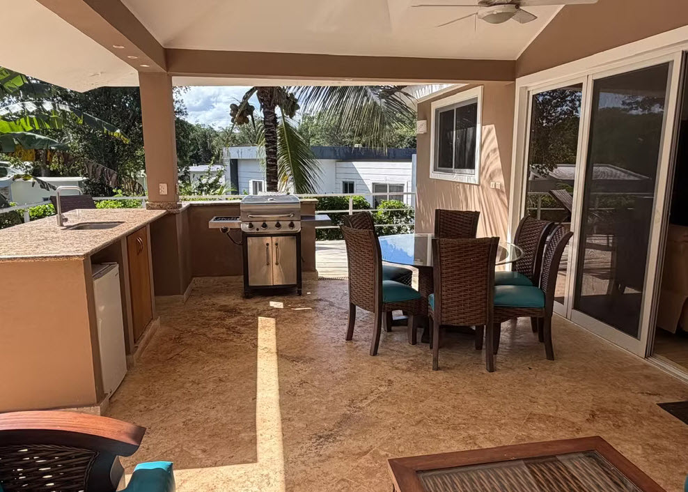 Covered patio with outdoor kitchen, grill, and dining set overlooking tropical foliage.