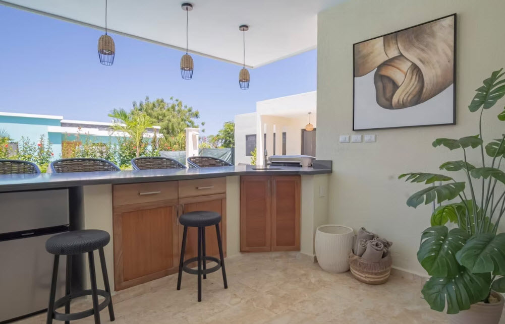 Modern outdoor kitchen with bar stools, mini-fridge, and tropical plants.