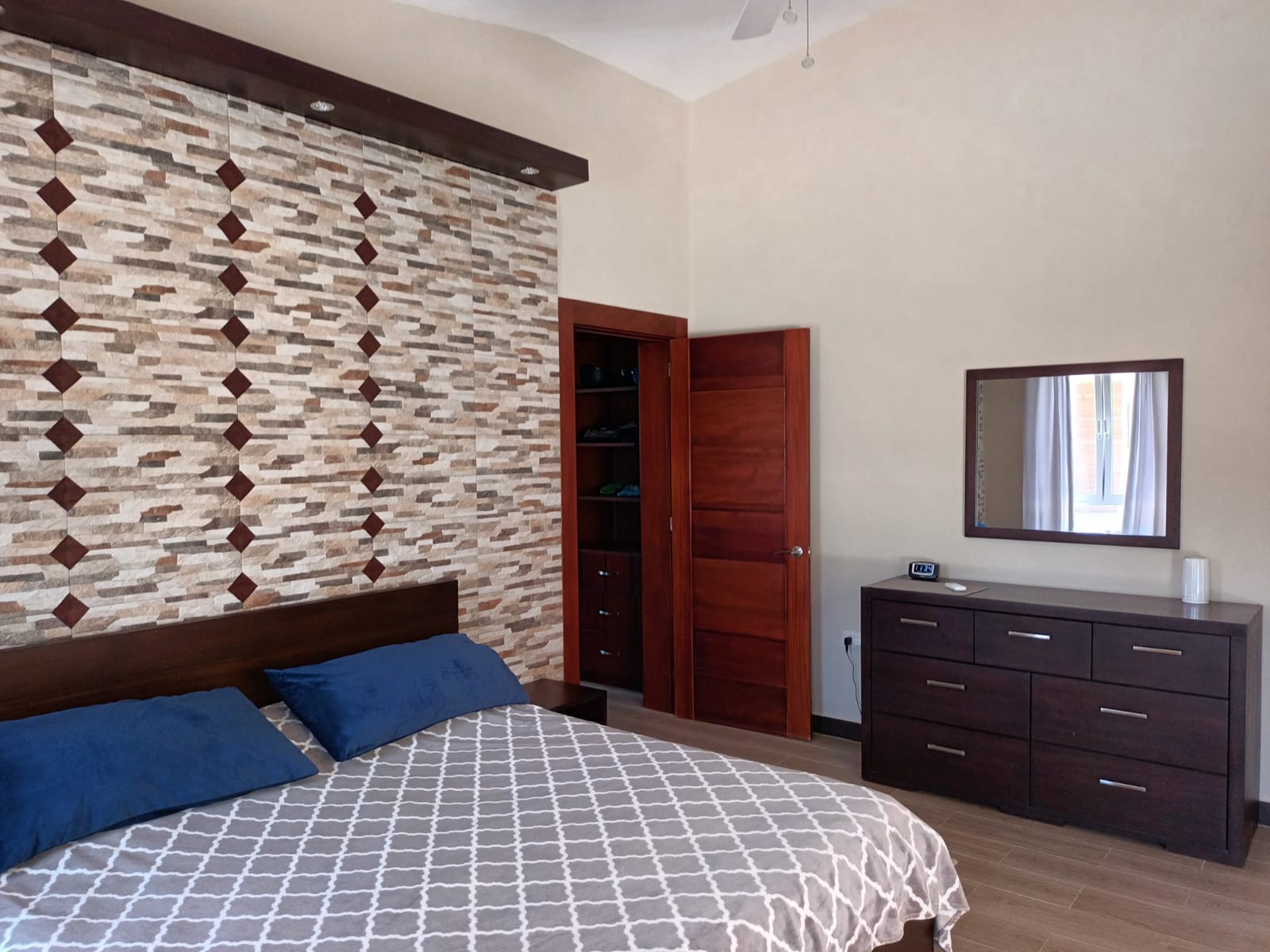 Modern bedroom with patterned accent wall, wooden closet, and dresser with mirror.