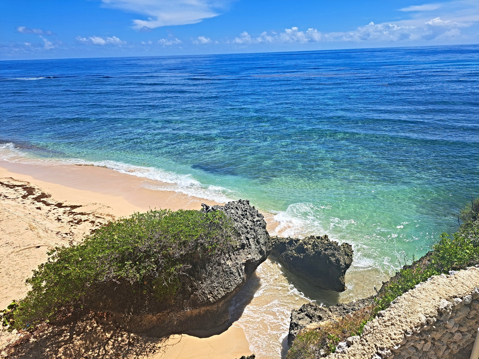 Crystal clear turquoise ocean water meets a sandy beach with rocky outcrops and lush green foliage.