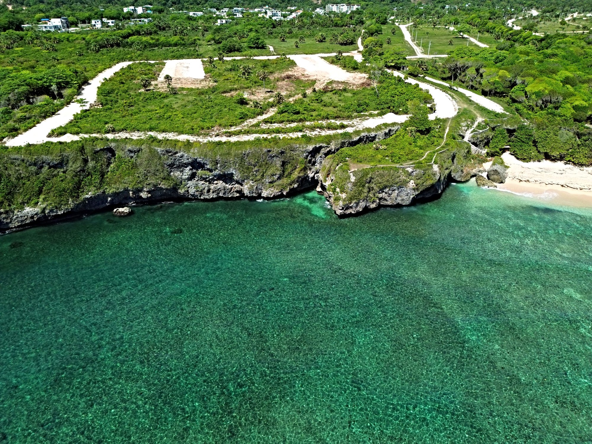 Aerial view of a coastal cliff with a small airstrip and lush green vegetation overlooking turquoise waters and a sandy beach.