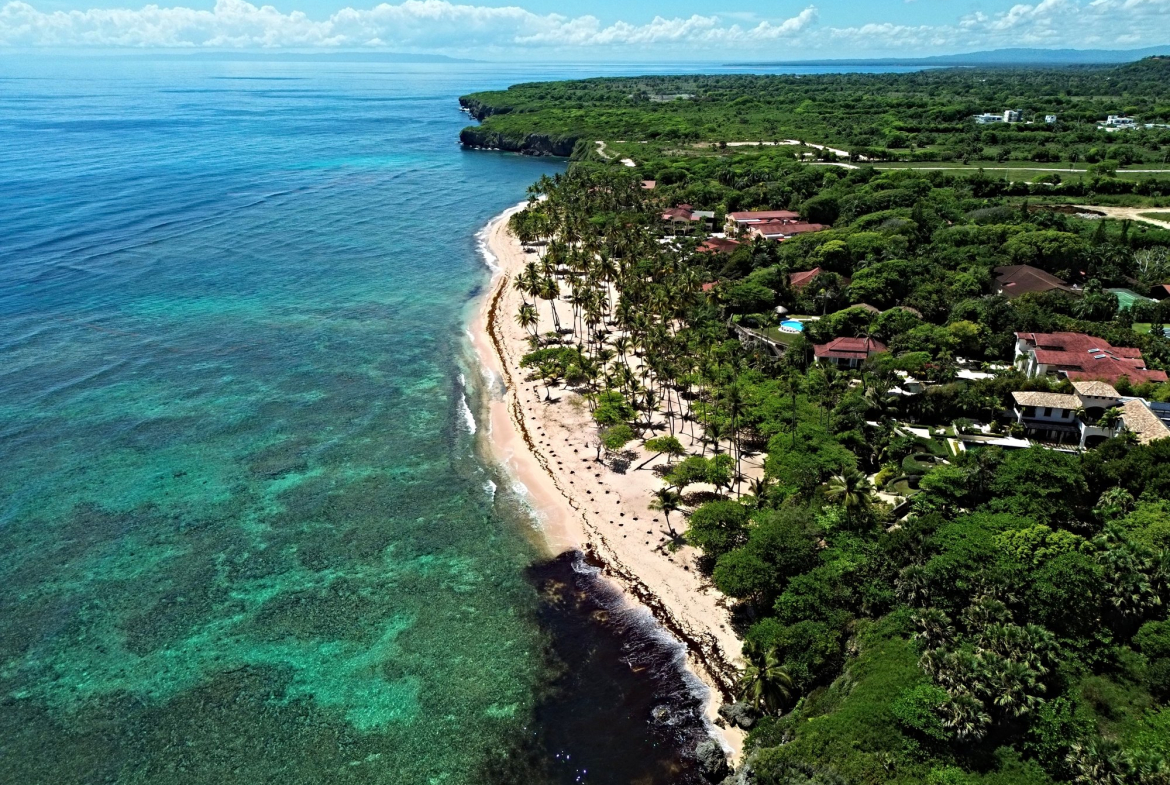 Aerial view of a tropical coastline with turquoise water, sandy beach, palm trees, and villas.