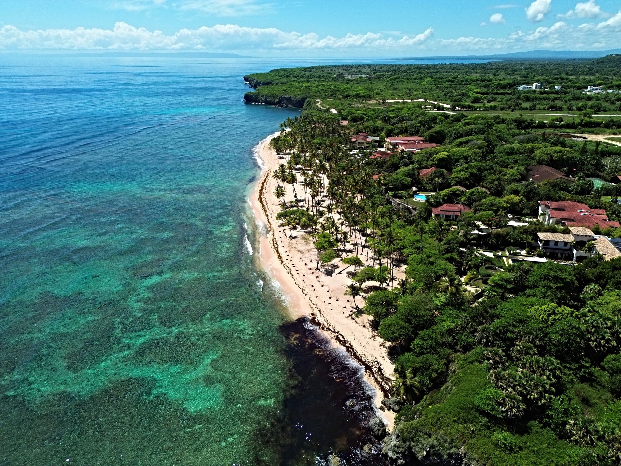 Aerial view of a tropical coastline with turquoise water, sandy beach, palm trees, and villas.