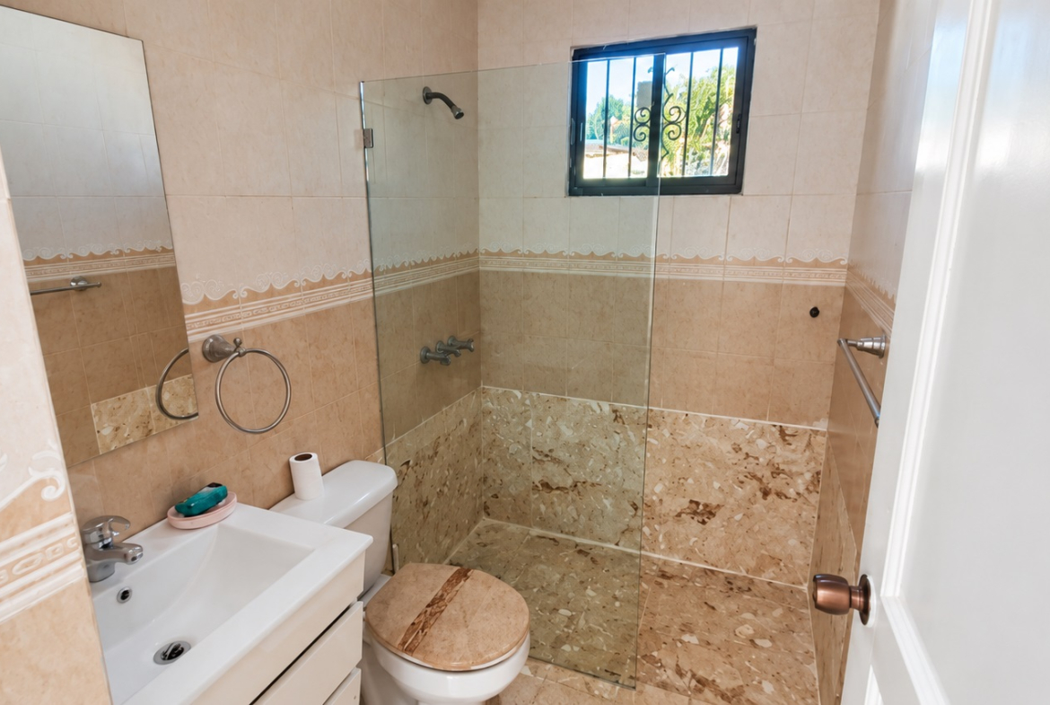 Small beige tiled bathroom with white sink, toilet, and glass shower.